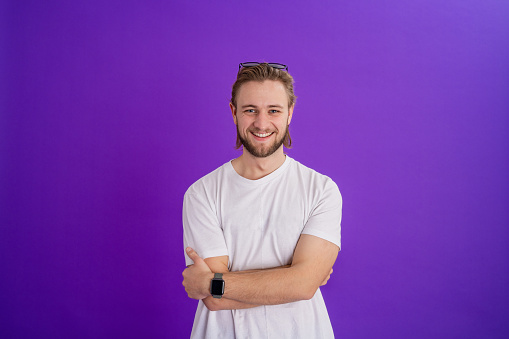 A studio portrait shot of a young man wearing a t-shirt standing in front of a purple backdrop while looking at the camera and smiling with his arms crossed.