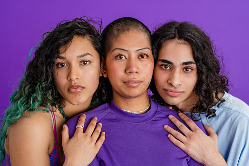 A studio portrait shot of a two female friends and their male friend, standing together in front of a purple backdrop. They are cheek to cheek with each other while looking at the camera with a contented look on their faces.