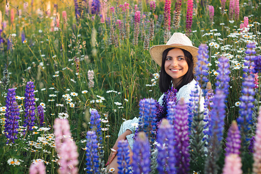 Portrait of young attractive woman in dress and straw hat holding bouquet of blooming lupin flowers on summer meadow at sunset.