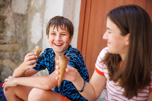 Preadolescent boy and his teenage sister sitting on the front door stairs and eating ice cream in cone on summer holiday