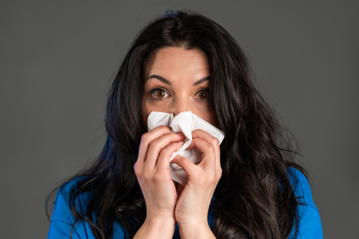 Mature woman sneezes into tissue. Isolated on grey studio background. Adult lady is sick, has a cold or allergic reaction. Coronavirus, epidemic 2020, illness concept