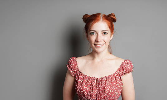 happy young woman with big toothy smile and red hair space buns on gray background with copy space