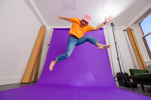A shot in a studio of a young man wearing a bucket hat and sunglasses jumping and leaping in front of a purple backdrop while looking at the camera and looking excited, his hands are outstretched while he jumps. Studio equipment is surrounding him.