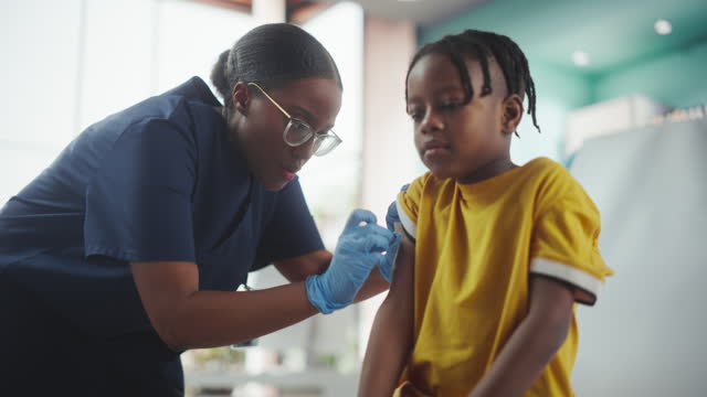 Young African American Boy Sitting In The Chair In Bright Hospital And Getting His Flu Vaccine. Female Black Nurse Is Performing Injection. Professional Woman High-Fives A Worried Kid For Being Brave.