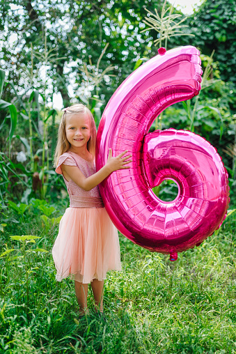 Happy girl holding a balloon on her sixth birthday in the backyard. Funny emotional child with pink balloon in the garden. 6 six years old, 6th birthday party celebration balloon.