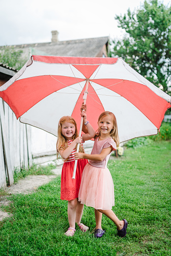 Happy funny childs with big beach umbrella under summer shower or heavy rain in the backyard. Girls walk is wearing dress and enjoying rainfall in spring park. Kids playing and catching rain drops.