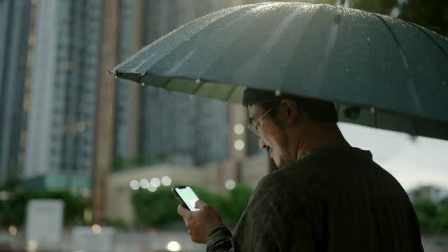Young man using a cellphone and holding an umbrella while walking outside in the rain