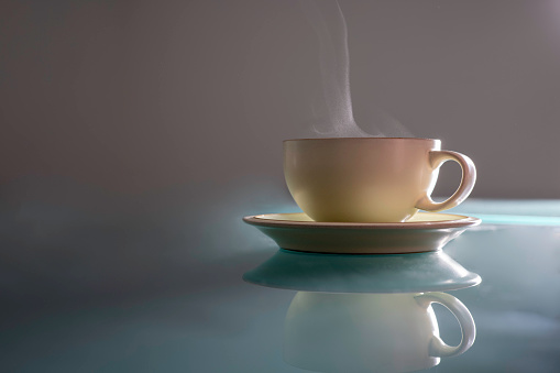 A side lit cup and saucer sit on a glass table, steam is visible rising from the cup.