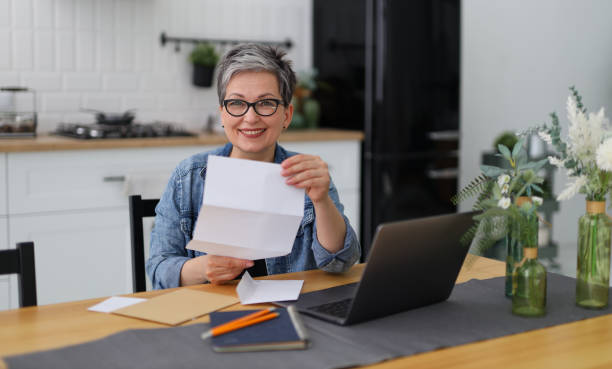 a woman joyfully reads a letter or invitation while sitting at table with a laptop. - news of the world imagens e fotografias de stock