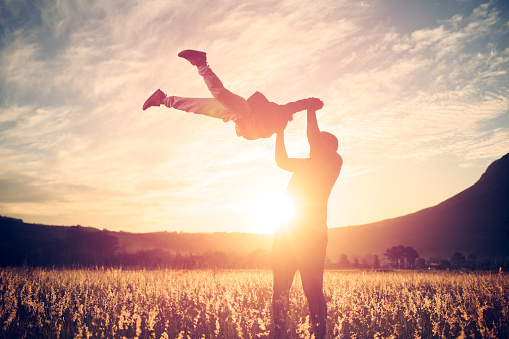Silhouette of a man swinging a child with legs wide open flying through the air holding hands at sunset in a golden field Stellenbosch, South Africa