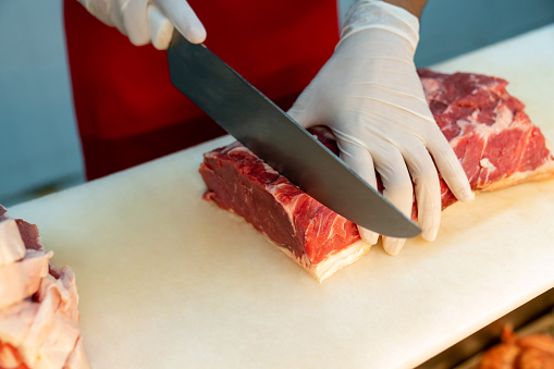 Close up shot of unrecognizable butcher wearing protective gloves cutting slices of meat at the butcher's shop - People at work concepts