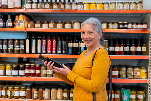 Portrait of senior woman at the supermarket holding a bottle of balsamic vinegar while facing the camera smiling - Lifestyles concepts