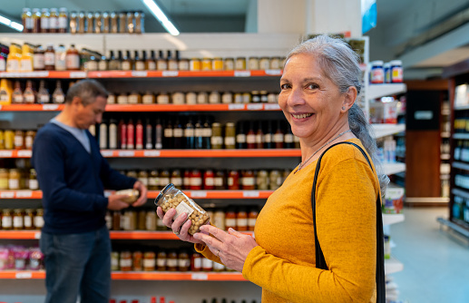 Portrait of senior woman at the supermarket holding a jar of chickpeas shopping for groceries with her husband while she smiles at the camera - Lifestyles concepts
