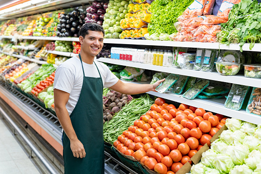 Cheerful salesman holding packaged vegetables at the refrigerated produce aisle in the supermarket while facing the camera smiling - Place of work concepts