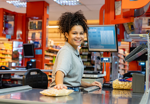 Portrait of cheerful young cashier facing the camera smiling while scanning products for a customer - People at work concepts