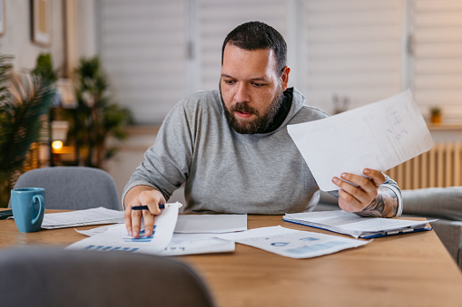 Man checking personal finances at home, while sitting at the table, holding the paper.