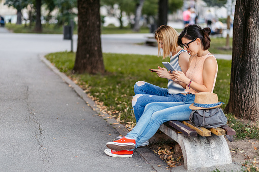 Two young women looking at their devices and sitting next to each other in the park.