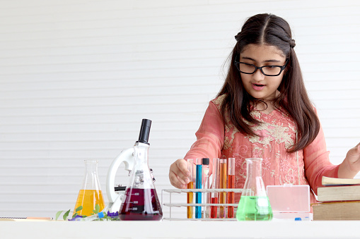 Cute Indian school girl in India traditional dress costume doing science experiments in laboratory, young scientist kid with microscope and lab equipment learning biologics and chemistry in classroom.