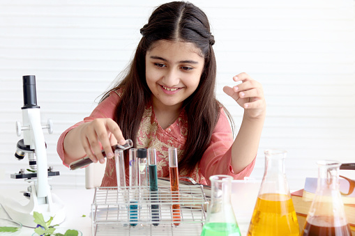 Cute Indian school girl in India traditional dress costume doing science experiments in laboratory, young scientist kid with microscope and lab equipment learning biologics and chemistry in classroom.