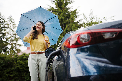 Woman calling, standing under umbrella while charging her electric car ona rainy day. Potential risks with charging an electric vehicle in the rain. Electric vehicle with charger in charging port.