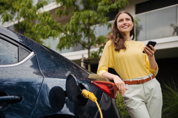 Young woman with smartphone waiting while her electric car charging in home charging station, sustainable and economic transportation concept. stock photo