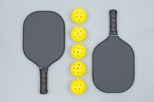 Flatlay close view of two black pickleball paddles with five yellow balls on light gray background