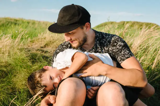 Photo of Father's Day. Happy son hugging dad and sitting on the green grass in the field in summer mountains. Dad and son playing at sunset. Kid with parents smiling, spending time together. Children's Day.