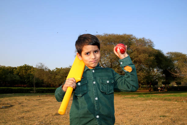 Young cricket player of Indian ethnicity standing portrait holding bat and ball in hands outdoor in park during sunset.