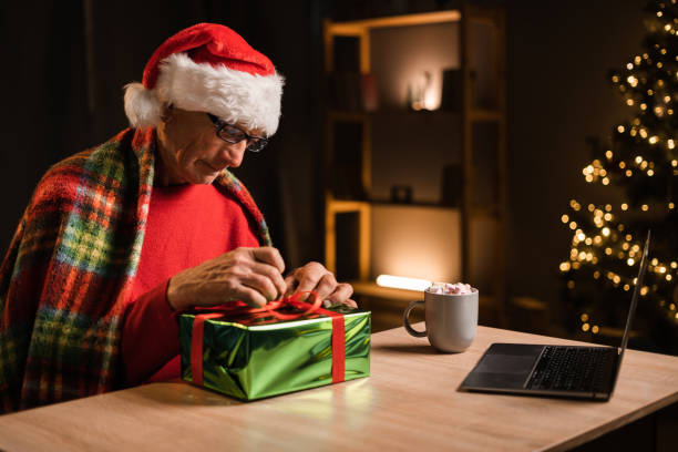 istock Happy old man wrapping gift sits at table in front of laptop has video call with relatives on christmas eve. Christmas celebration 1497206909