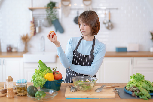 Asian woman looking at fresh tomatoes in hand and thinking what to cook, Happy Asian woman cooking healthy food in the kitchen.
