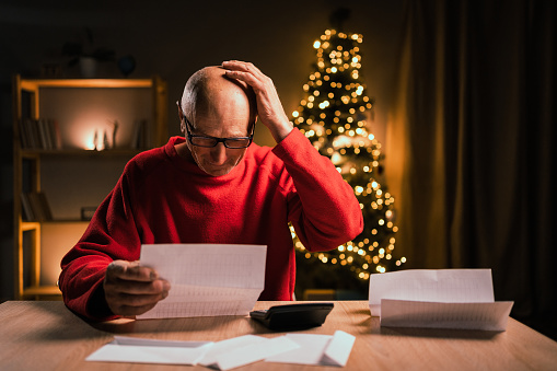 Christmas stress. Old frustrated man holding many bills counting expenses for winter holidays. Calculator on the table, Christmas decoration over background. Copy space