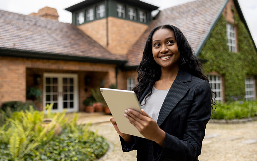 Portrait of a happy African American real estate agent using her tablet in front of a house for sale