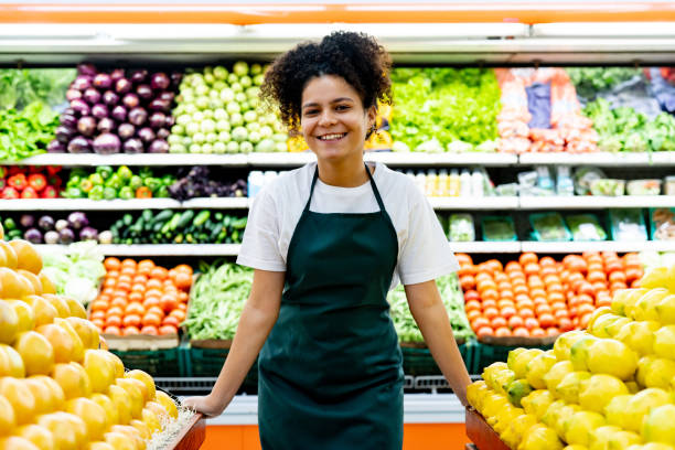istock Latin American young woman working at a supermarket leaning hands on fruit display while facing camera smiling very cheerfully 1497211501