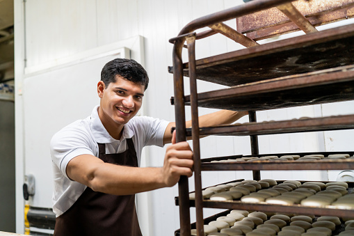 Portrait of Latin American man working at a bakery pushing trays of cookies to the oven while facing the camera smiling - People at work concepts