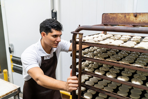 Latin American male baker pushing industrial trays with cookies to the oven  - People at work concepts