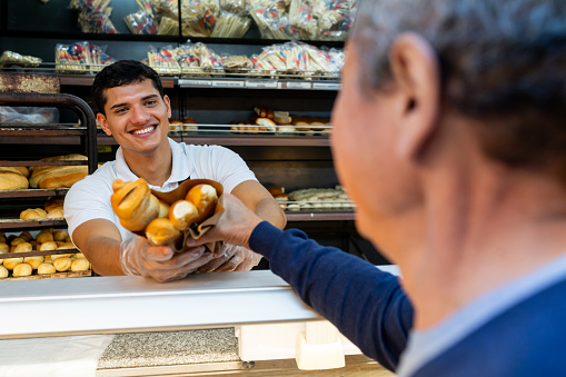 Friendly salesclerk handing a bag of bread to unrecognizable customer at a bakery - People at  work concepts