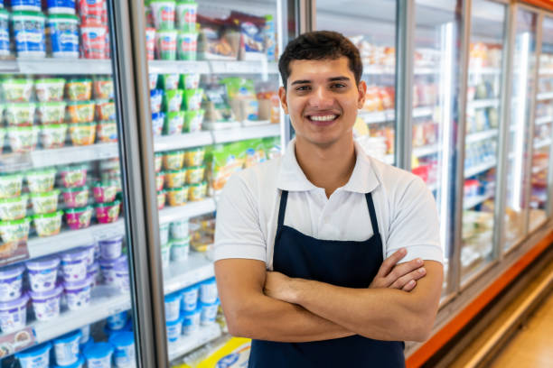 portrait d’un jeune homme travaillant au supermarché dans la section réfrigérée face à la caméra souriant les bras croisés - grocer photos et images de collection
