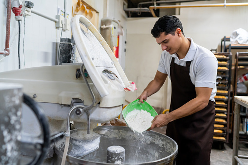 Young male baker adding flour to an industrial mixer to prepare bread at a factory