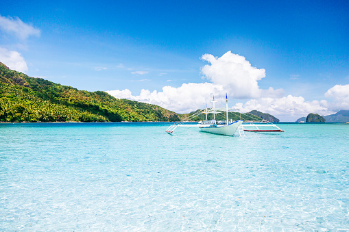 Tropical island in the Philippines on a sunny day. In the foreground, a traditional Filipino white boat takes center stage, peacefully floating in the pristine waters of a crystal clear sea lagoon. In the distance, the island reveals its scenic beauty with lush hills adorned in abundant tropical plants. The vibrant blue sky above is adorned with fluffy clouds, completing the picturesque scene.