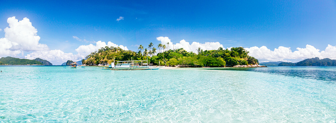 Panoramic view of Snake Island in the Philippines on a sunny day. The foreground showcases the transparent and turquoise sea, while the island itself is visible in the distance. The island is characterized by its hills covered in lush tropical plants and palm tree groves. A white beach can also be seen on the island. Near the island, there are several traditional Filipino white boats. The sky above is vibrant blue with fluffy clouds, and there are additional island silhouettes in the background.