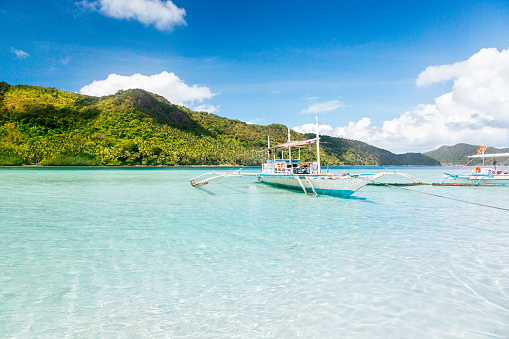 Tropical island in the Philippines on a sunny day. In the foreground, there is a traditional Filipino white boat peacefully floating on the calm waters of a crystal clear sea lagoon. The island's landscape is characterized by lush hills covered in abundant tropical plants. The sky above is blue with scattered fluffy clouds.
