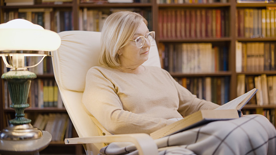 Woman in her 50s reading a book late in the evening in her home library, relaxing time