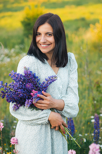 Portrait of young attractive woman in a dress, holding bouquet of blooming lupin flowers on summer meadow at sunset.
