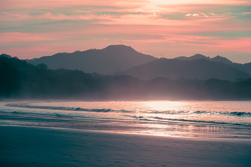 The sun rises behind the dry forests in Playa Carrillo, Guanacaste Province of Costa Rica. The ocean mist is illuminated in red light from the rising sun.