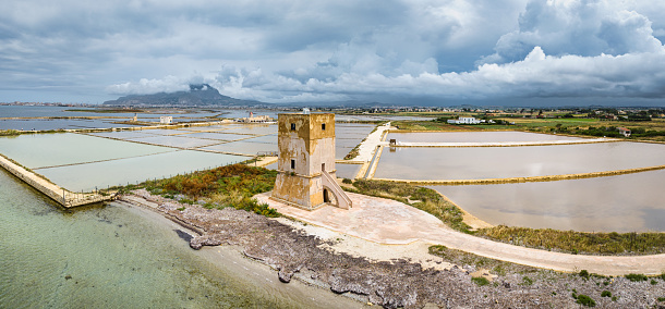 Panorama of ancient Saltworks Torre Nubia under dramatic moody skyscape at Saline di Trapani - ancient Saltworks. Drone point of view over the salt works - saline ponds at the western coast of Sicily Island. Ancient Saline Trapani, Marsala, Western Coast Sicily Island, Southern Italy, Southern Europe.