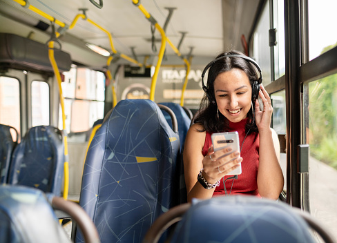 Happy young woman wearing earphones and listening to music on a smartphone while sitting alone on a bus