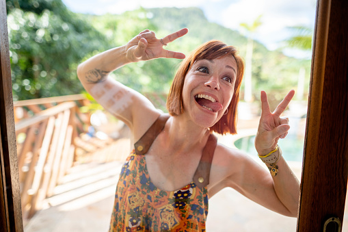 Funny mature woman standing outside and looking inside from window sticking out tongue and making peace signs with her hands