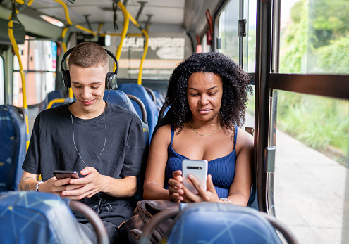 Two young friends streaming videos on their smart phones while riding together on a city bus