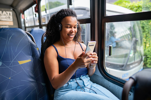 Smiling young woman wearing headphones streaming a video on her smart phone while riding on a bus