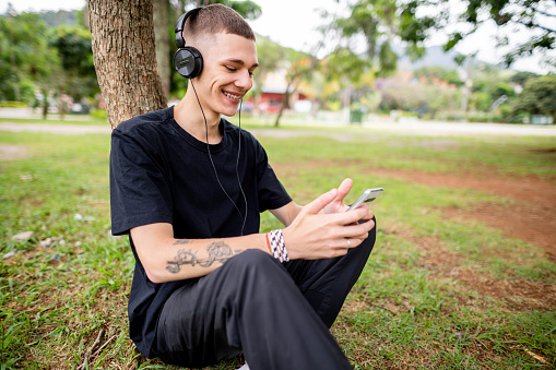 Smiling young man wearing headphones leaning against a tree in a park and streaming a video on his smart phone